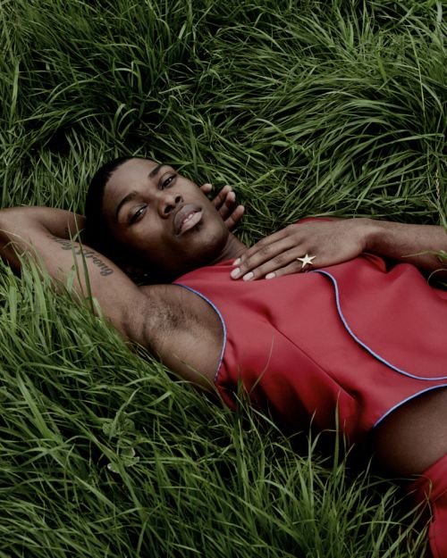 An African American adult male with a star ring, wearing a red sleeveless shirt, laying in green grass in a close-up shot.