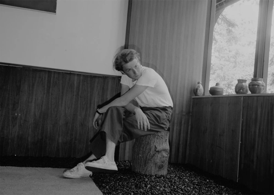 Young man poses in relaxed indoor shot. He's seated on a wood stump with gravel floor and wood paneled walls. Black and white.