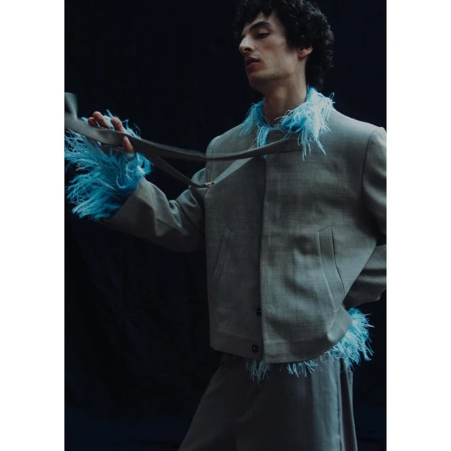 An androgynous model poses in a grey feather-trimmed jacket and matching skirt against a dark background, showcasing unique fashion.