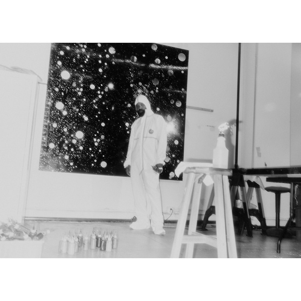 Artist in white suit stands next to large black painting with white dots inside a studio, surrounded by art supplies and tools.