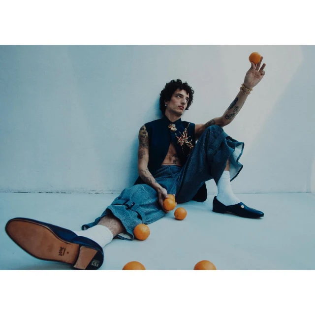 Adult male model sits in a studio setting with oranges wearing casual pants, dark sleeveless top, and loafers with white socks.