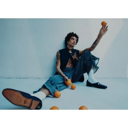 Adult male model sits in a studio setting with oranges wearing casual pants, dark sleeveless top, and loafers with white socks.
