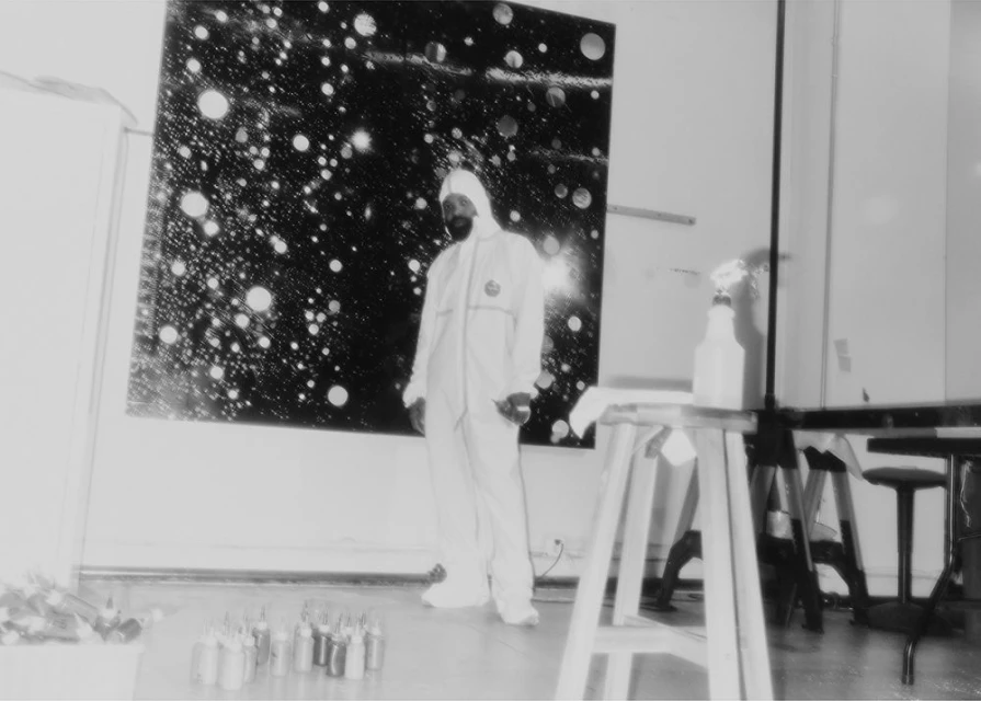 Artist in paint suit stands before their abstract cosmic art piece, featuring spray cans, tables and chairs, captured in a black and white studio.