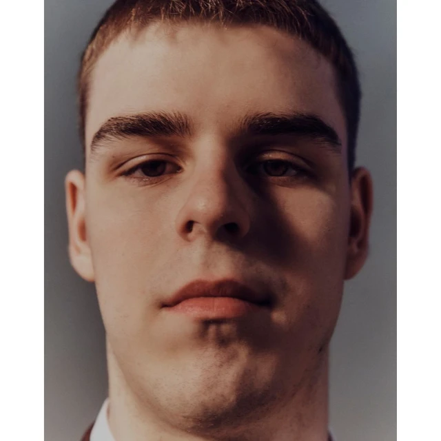 Close-up portrait of a young adult male with short brown hair, fair skin, and brown eyes, wearing a brown jacket against a grey background.