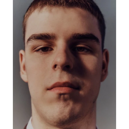 Close-up portrait of a young adult male with short brown hair, fair skin, and brown eyes, wearing a brown jacket against a grey background.