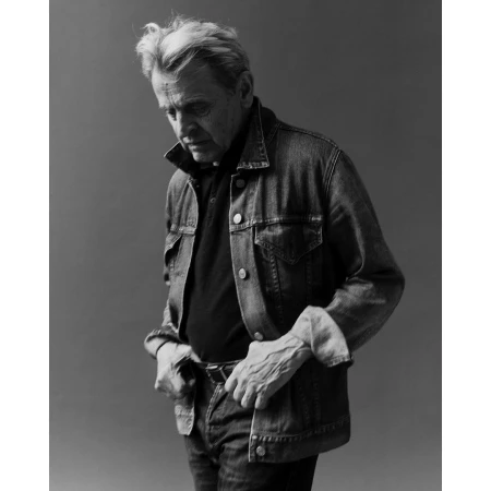 Monochrome portrait of an elderly man in a denim jacket and jeans, looking down with his hands near his waist.