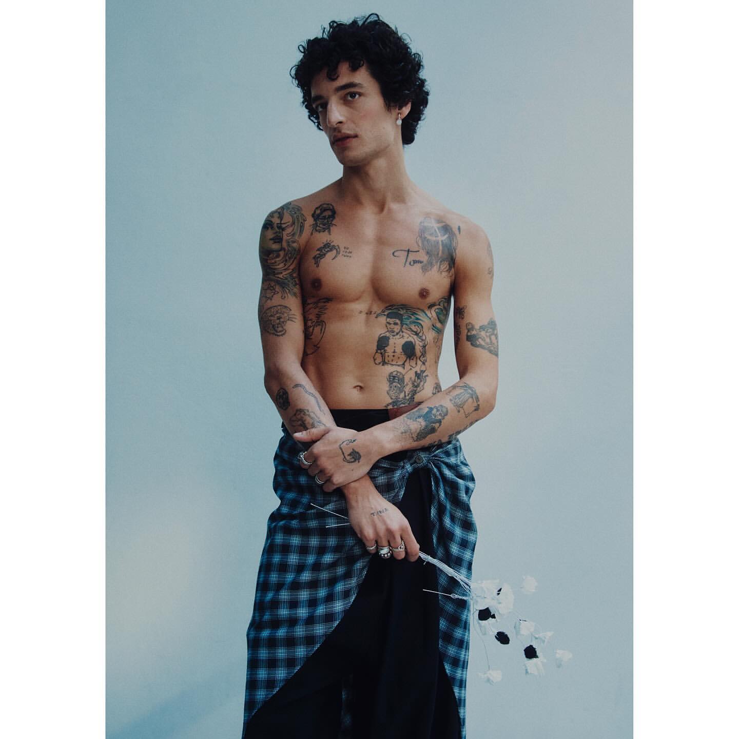 Adult male with curly hair and tattoos poses shirtless indoors holding white flowers, wearing a plaid wrap skirt and black underpants.