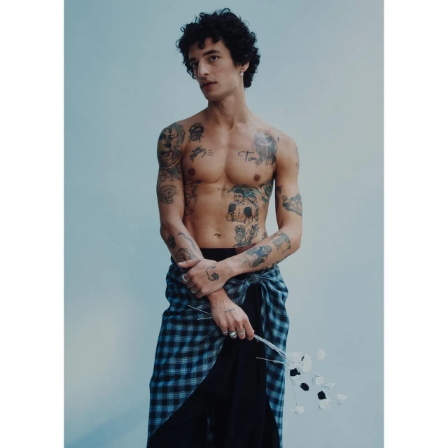 Adult male with curly hair and tattoos poses shirtless indoors holding white flowers, wearing a plaid wrap skirt and black underpants.