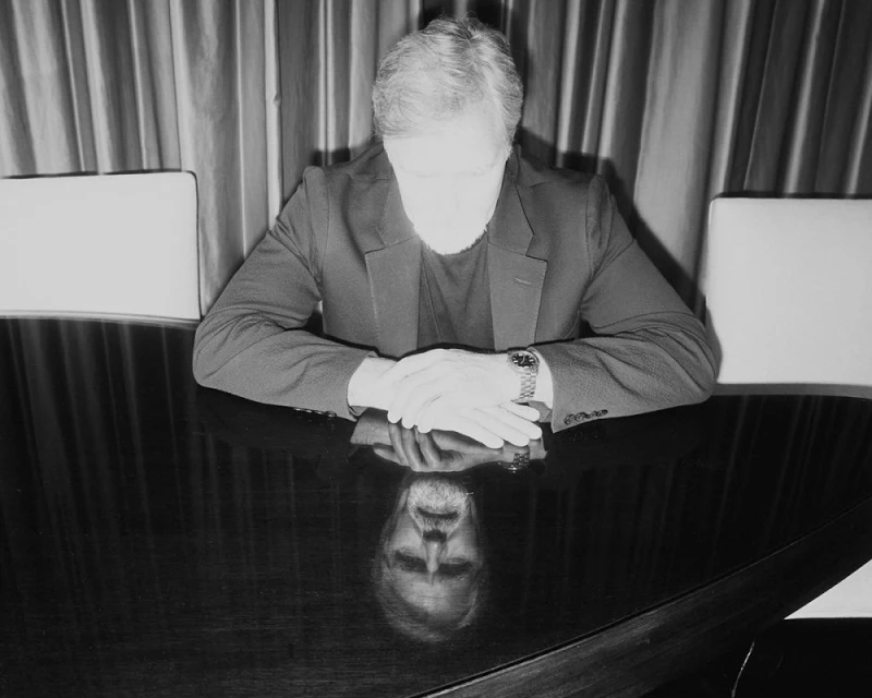 Monochrome portrait of a seated adult male with light hair wearing a suit, reflecting on a dark table in an indoor setting.