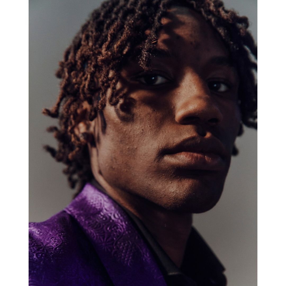 Portrait of a young black male with dreadlocks in close-up, wearing a purple patterned suit jacket in a photo studio setting.