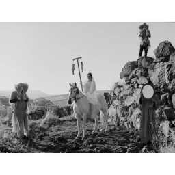 Black and white editorial photograph featuring a woman on a white horse surrounded by figures holding props in a rocky landscape.