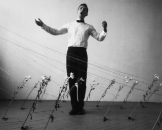 Man in white dress shirt and black trousers with bow tie stands amidst strings attached to tilted flowers.