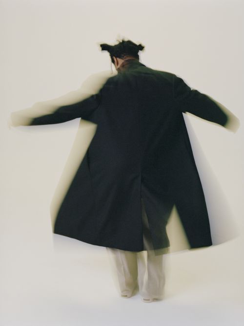 Fashion shot of a person in motion wearing a long dark coat and light pants against a white background in a photo studio.