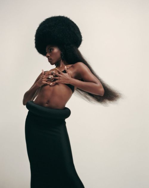 Woman with large afro poses in black skirt and bra top against light background; hands near chest, side view.
