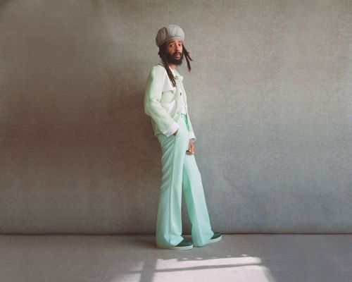 An adult male poses in a light green suit with bell-bottom pants and a cream-colored hat against a neutral-toned wall in a studio shot.
