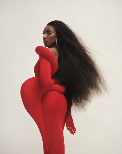 A woman with long dark hair poses in a red, full-body garment in a photo studio, creating a surreal and stylish portrait against a white backdrop.