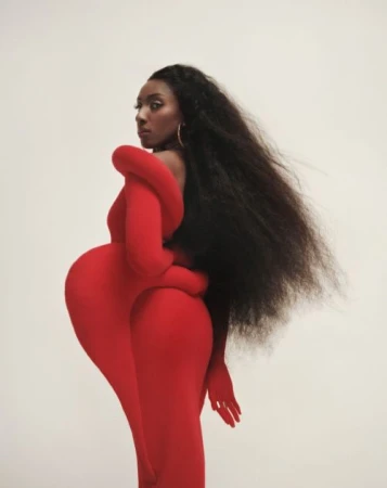 Black woman with long flowing dark hair wearing a vibrant red avant-garde sculptural dress against an off-white background.