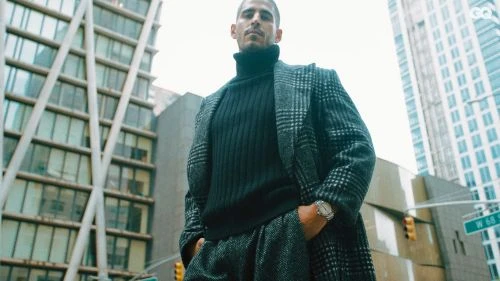 Man in coat and turtleneck stands in city street with buildings in background, wearing a watch and gazing forward in a low-angle shot.