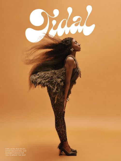 A woman poses on an ochre background. She wears a sparkling brown catsuit and feathered top, with hair blowing behind her head.