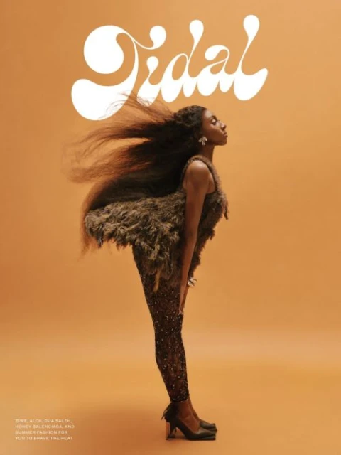 A woman poses on an ochre background. She wears a sparkling brown catsuit and feathered top, with hair blowing behind her head.