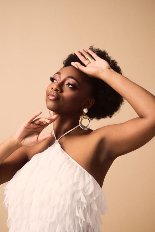 Young Black woman poses gracefully in a white, feathered top with large earrings and a ring against a tan background in a studio setting.