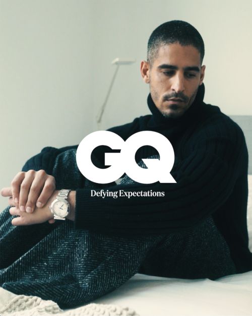 Man in a black turtleneck sweater and tweed trousers seated on a bed wearing a silver wristwatch