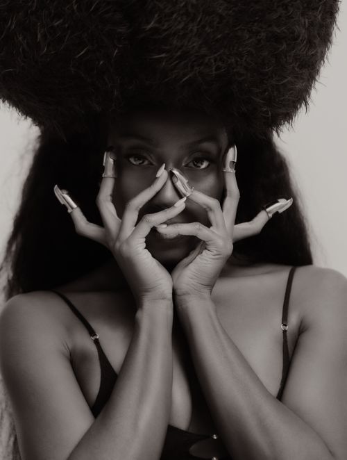 Portrait of a Black woman holding her hands to her face with silver claw rings, wearing a large textured afro hat against a plain background
