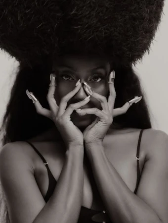 Portrait of a Black woman holding her hands to her face with silver claw rings, wearing a large textured afro hat against a plain background