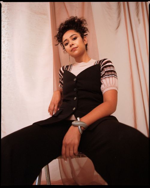 Portrait of a young woman with curly hair wearing a black vest, seated indoors against a light pink backdrop with sheer fabric.
