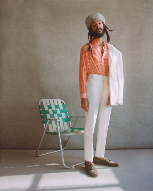 Adult male in an orange striped shirt and white suit with a grey hat posing next to a woven lawn chair indoors with a neutral background.