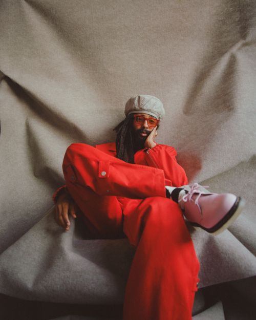 Portrait of a black man with a beard wearing a red suit and orange glasses, sitting on a wrinkled grey fabric backdrop, studio shot.