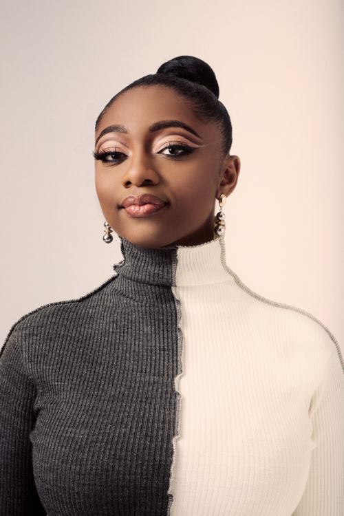 Portrait of a young woman with dark hair in a high bun, wearing a grey and white turtleneck sweater, against a neutral backdrop.