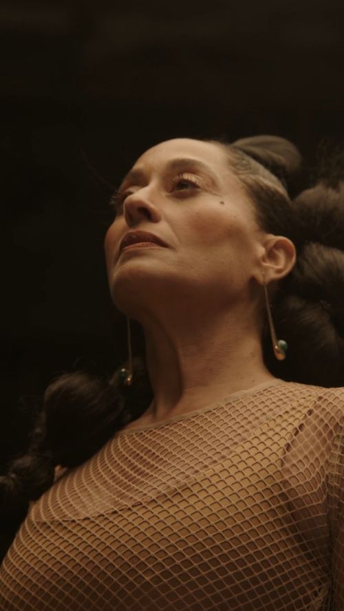 Elegant woman with braided hair and drop earrings in a close-up shot, wearing a net-patterned top in a dimly lit setting.