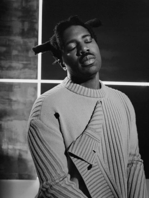 Black and white portrait of an adult male with locs and a sweater standing indoors with a geometric background.