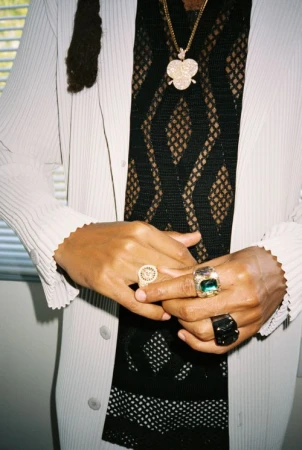 Close-up of a person wearing a white pleated coat, patterned black mesh top, gold pendant necklace, and several rings.