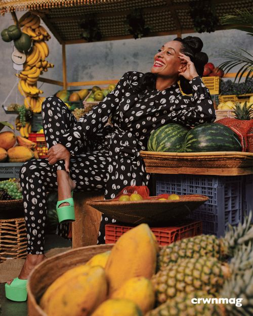 Elegant woman in patterned jumpsuit poses amidst a colorful fruit display, showcasing a vibrant fashion editorial with tropical flair.