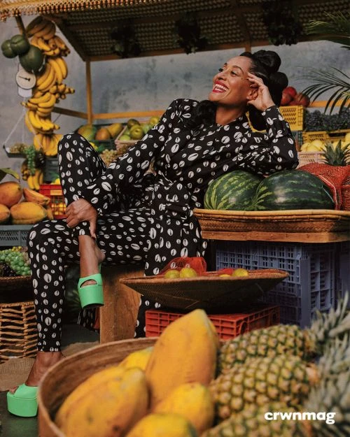Elegant woman in patterned jumpsuit poses amidst a colorful fruit display, showcasing a vibrant fashion editorial with tropical flair.