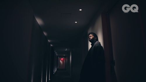 Adult man poses in dark hotel hallway wearing black coat. Light from end of corridor illuminates figure; GQ magazine logo visible.