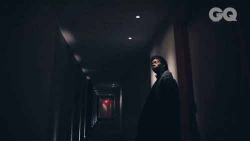 Adult man poses in dark hotel hallway wearing black coat. Light from end of corridor illuminates figure; GQ magazine logo visible.