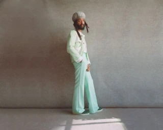 Man with dreadlocks wearing a light green suit and grey beret standing against a textured grey backdrop