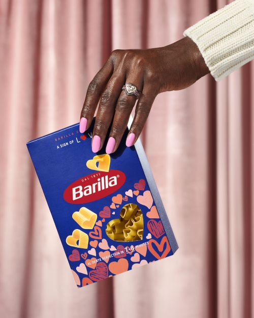 A hand with pink nails and a ring holding a box of Barilla heart-shaped pasta against a background of pink curtains.