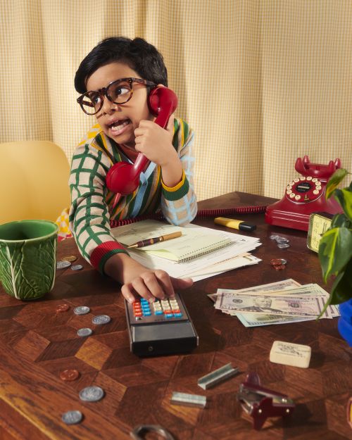 Young person with glasses sits at a desk using an old calculator while talking on a red rotary phone, surrounded by office props.