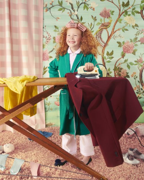 A young girl with red curly hair wearing a robe ironing on an ironing board in front of floral wallpaper and pink checkered curtains.