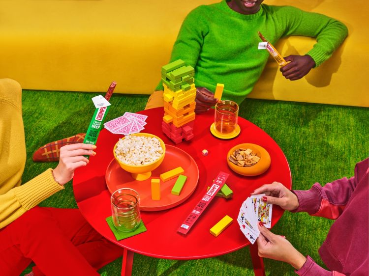 Diverse group playing games around a red table, enjoying snacks and drinks with a vibrant, colorful backdrop and cheerful atmosphere.