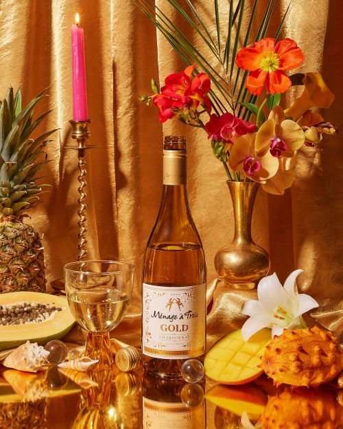 Wine bottle and glass with tropical fruits and flowers on golden reflective surface in studio lighting creating a vibrant still life.