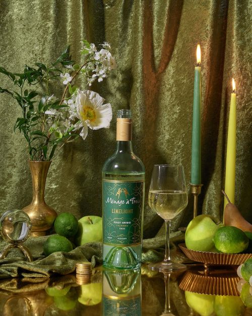 Wine bottle and glass of white wine on a reflective surface, with limes, green apples, and white flowers against a green velvet background.