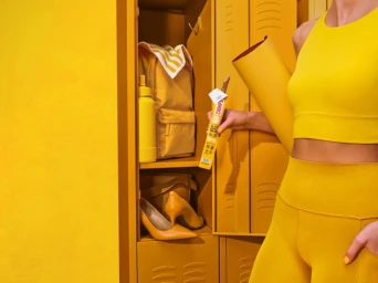 Woman wearing yellow athletic wear holding a snack stick and yoga mat standing beside yellow lockers.