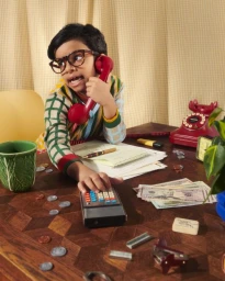 Child wearing thick glasses and a patterned sweater holding a red telephone receiver and operating a calculator at a wooden desk.