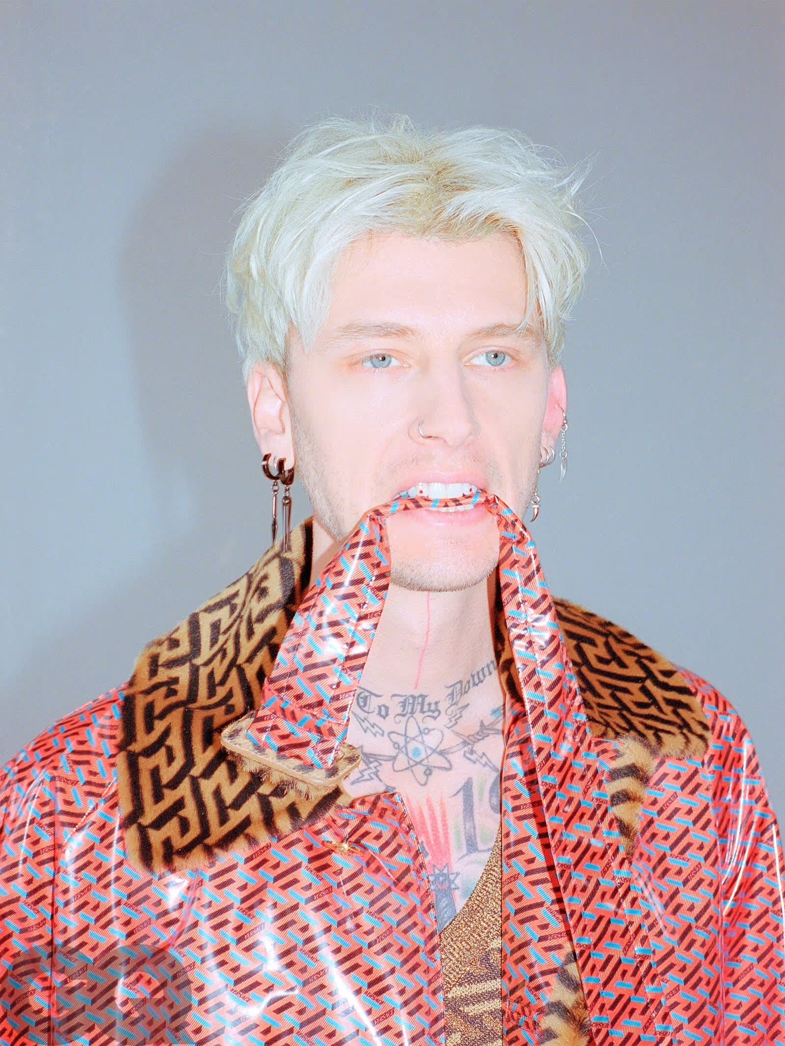 Portrait of a male celebrity with platinum hair wearing an patterned coat and holding the collar with his teeth in an indoor setting.