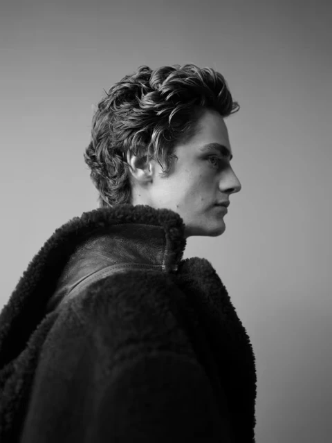 Monochrome portrait of a young adult male with curly hair wearing a shearling jacket, in a side profile view against a plain background.